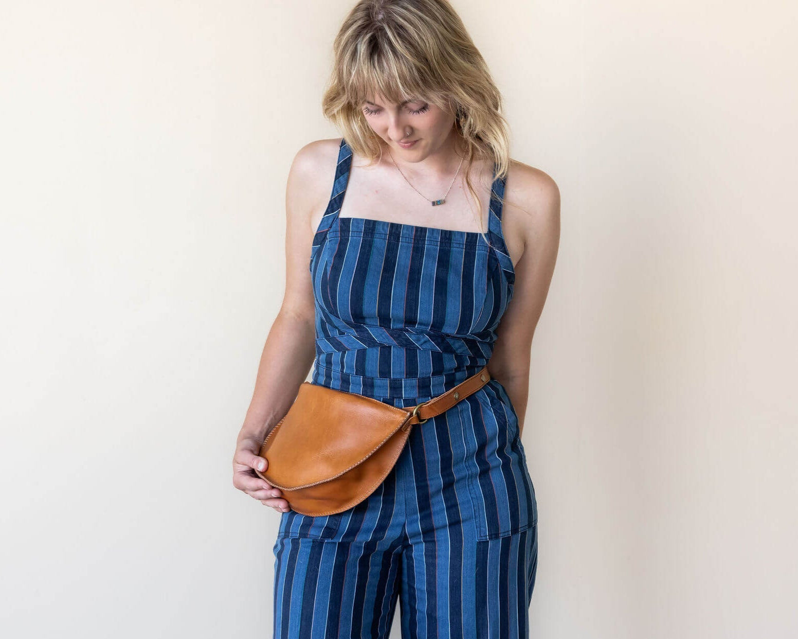 Woman wearing a blue striped jumpsuit with a brown leather belt bag against a plain background