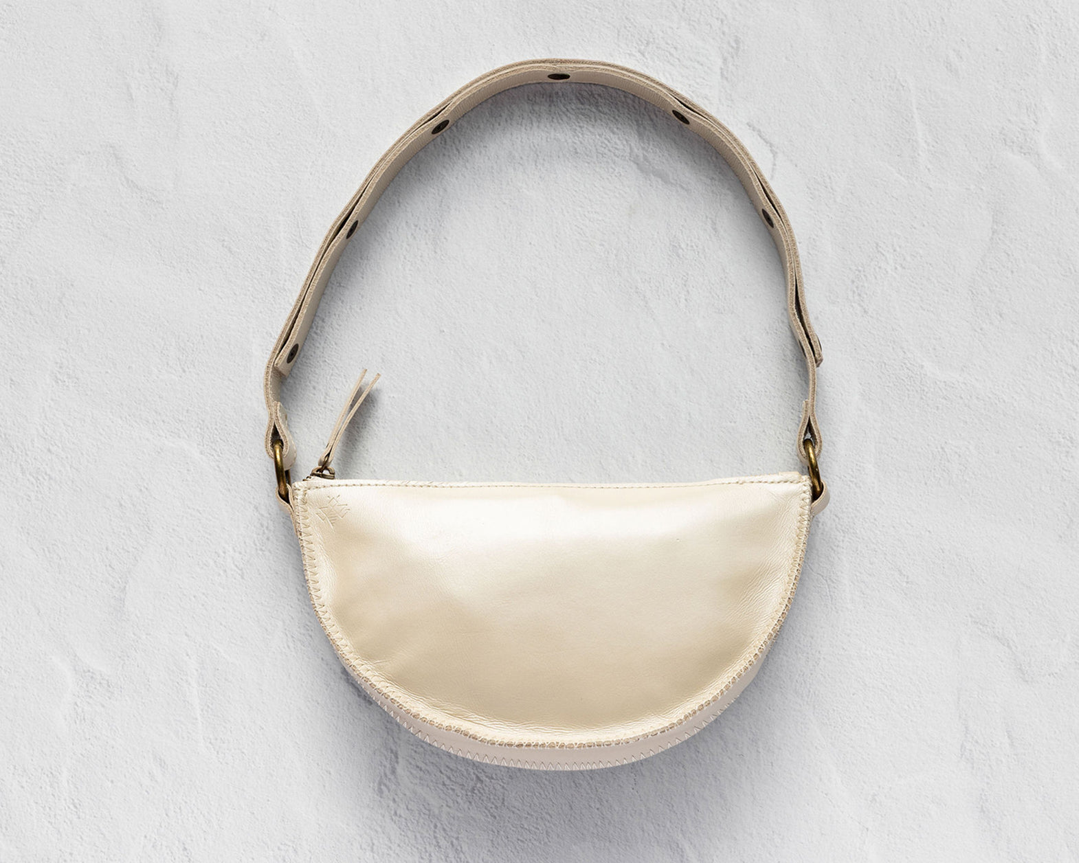 A cream-colored faux leather crescent-shaped shoulder bag with a matching shoulder strap, gold-tone hardware, and a small tassel detail, displayed on a textured light gray background.