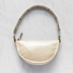 A cream-colored faux leather crescent-shaped shoulder bag with a matching shoulder strap, gold-tone hardware, and a small tassel detail, displayed on a textured light gray background.