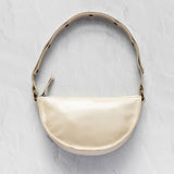 A cream-colored faux leather crescent-shaped shoulder bag with a matching shoulder strap, gold-tone hardware, and a small tassel detail, displayed on a textured light gray background.