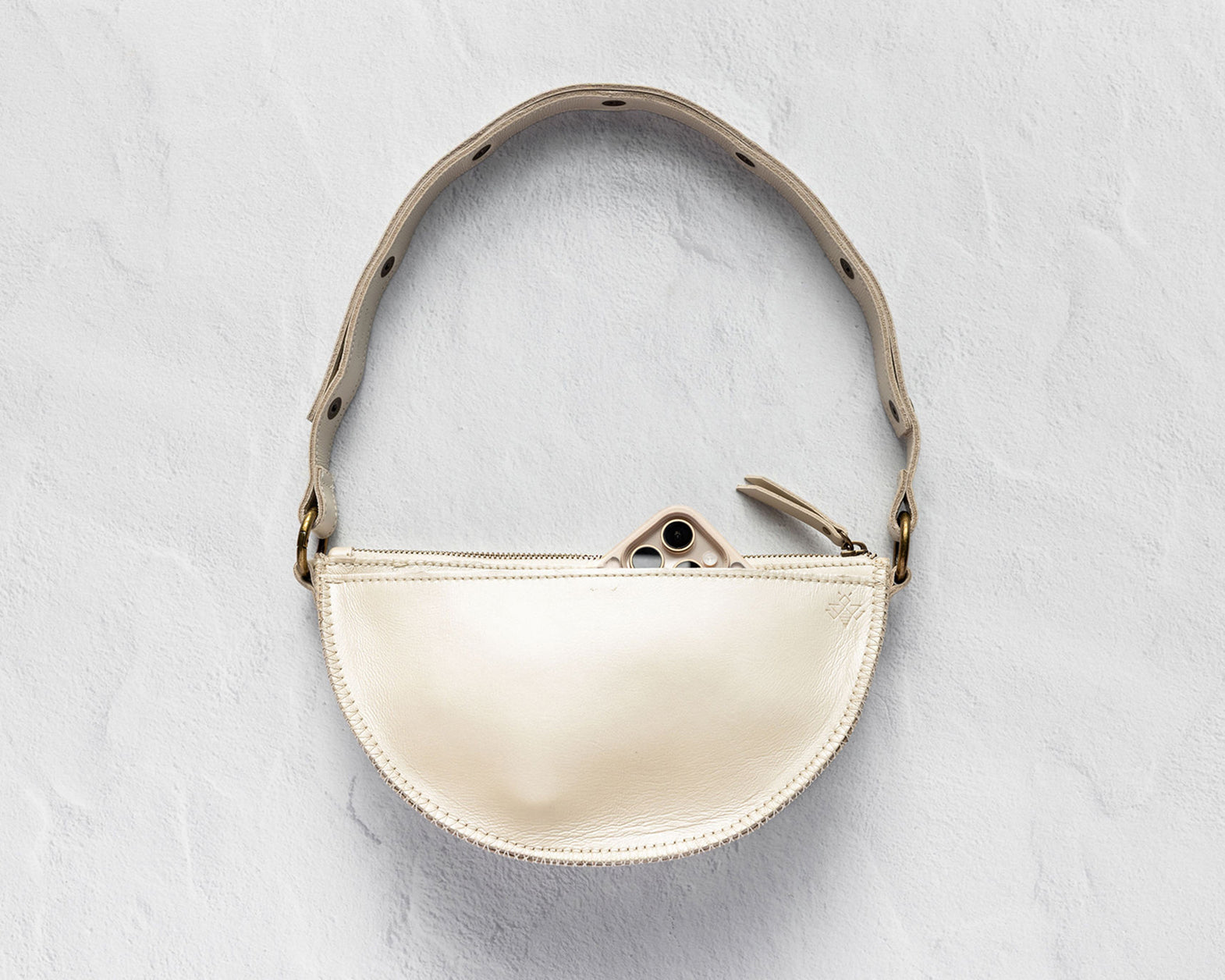 A small semi-circular cream leather shoulder bag with a matching leather strap and gold-tone hardware, displayed against a textured light gray background.