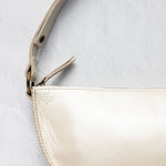 Close-up of a cream-colored leather shoulder bag with a matching leather strap and gold-tone hardware, set against a textured light gray background.