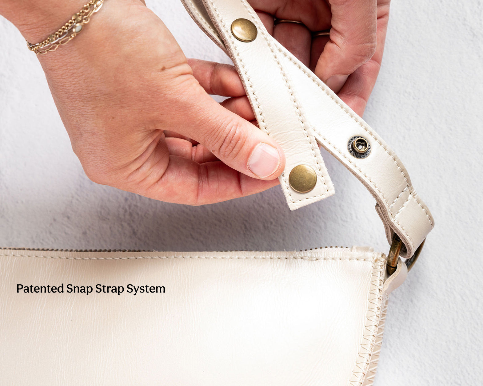 Close-up of a person's hands demonstrating the patented snap strap system on a cream-colored handbag. The strap is shown attaching to the bag with brass snap buttons.