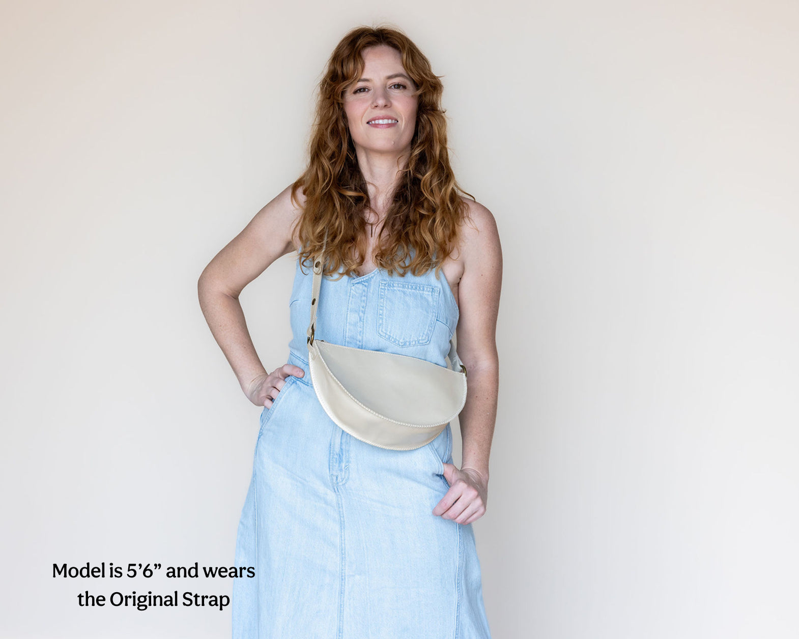 A woman with long wavy red hair models a light blue denim dress and a cream-colored crescent-shaped shoulder bag. The image has a text overlay in the bottom-left corner that reads 'Model is 5'6" and wears the Original Strap'.