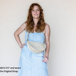 A woman with long wavy red hair models a light blue denim dress and a cream-colored crescent-shaped shoulder bag. The image has a text overlay in the bottom-left corner that reads 'Model is 5'6" and wears the Original Strap'.