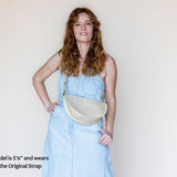 A woman with long wavy red hair models a light blue denim dress and a cream-colored crescent-shaped shoulder bag. The image has a text overlay in the bottom-left corner that reads 'Model is 5'6" and wears the Original Strap'.