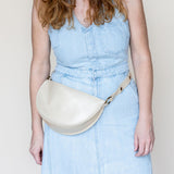 A woman wearing a light blue denim jumpsuit models a cream-colored faux leather half-moon fanny pack with a matching strap and gold-tone hardware against a plain off-white background.