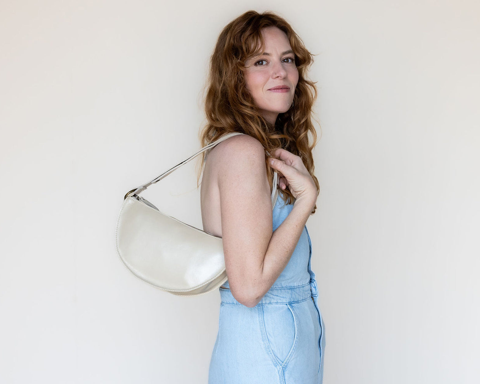 A woman with wavy light brown hair wearing a light blue denim jumpsuit and holding a cream-colored crescent-shaped shoulder bag against a plain off-white background.