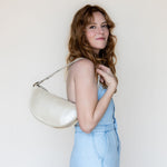 A woman with wavy light brown hair wearing a light blue denim jumpsuit and holding a cream-colored crescent-shaped shoulder bag against a plain off-white background.