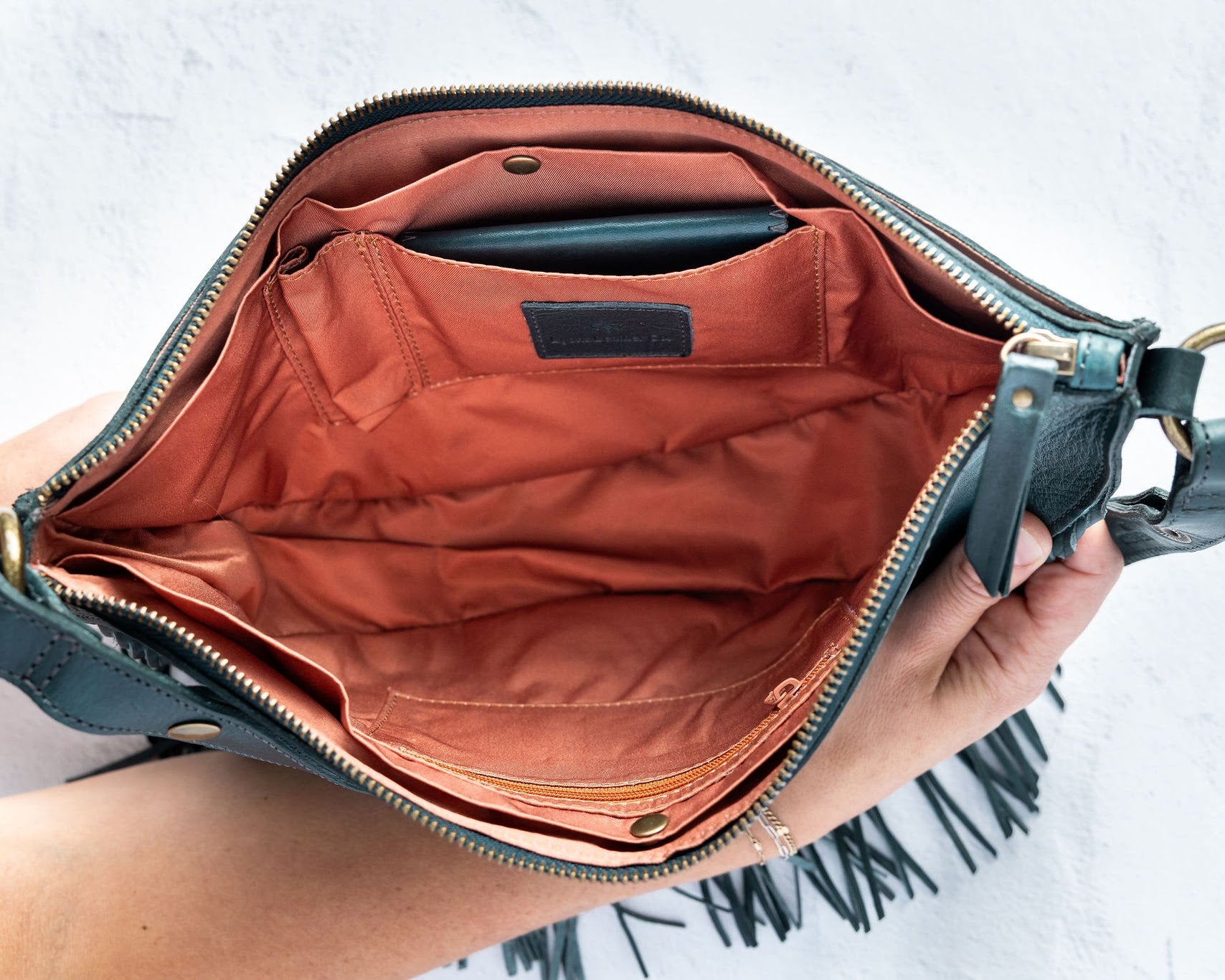 Teal leather fringe bag held open by a hand, showing the Pecan lining, on a white background