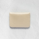 A minimalist cream-colored leather card holder or wallet with visible stitching and a debossed logo, displayed on a textured white background.