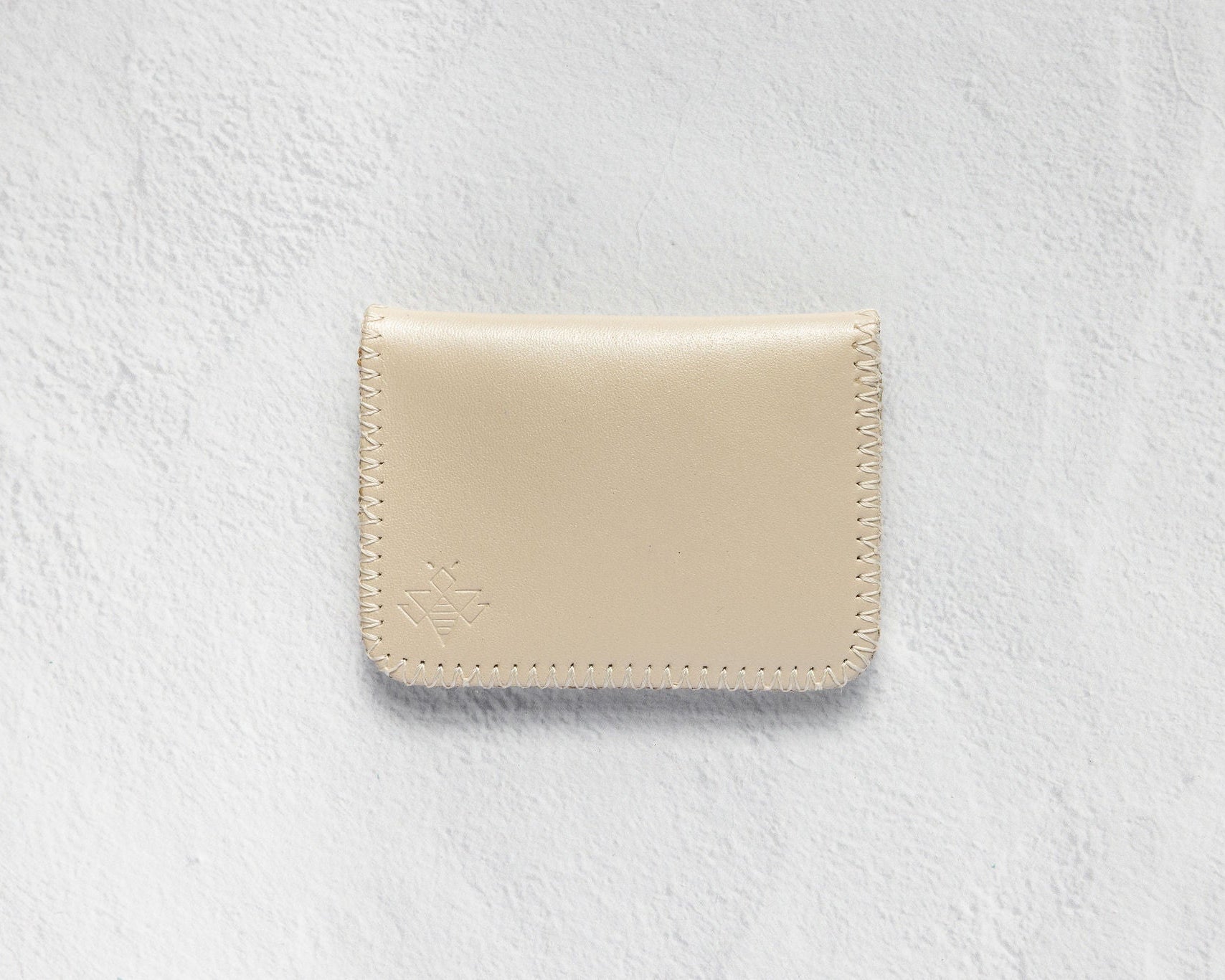 A minimalist cream-colored leather card holder or wallet with visible stitching and a debossed logo, displayed on a textured white background.