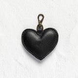 A black textured leather heart-shaped keychain with a brass clasp, displayed on a white textured background.