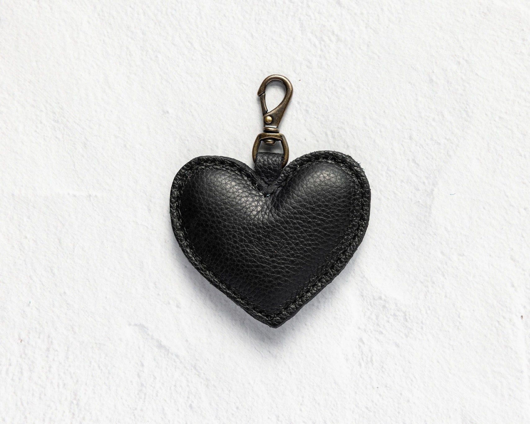A black textured leather heart-shaped keychain with a brass clasp, displayed on a white textured background.