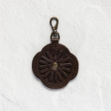 A dark brown leather keychain or bag charm with an embossed daisy flower design and a brass-colored lobster clasp, displayed on a textured white background.