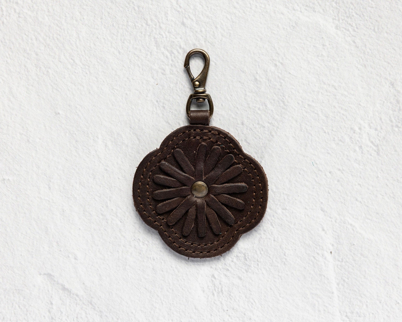A dark brown leather keychain or bag charm with an embossed daisy flower design and a brass-colored lobster clasp, displayed on a textured white background.
