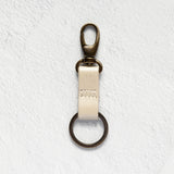 A brass key ring clasp with a cream-colored leather strap, shown against a textured white background.