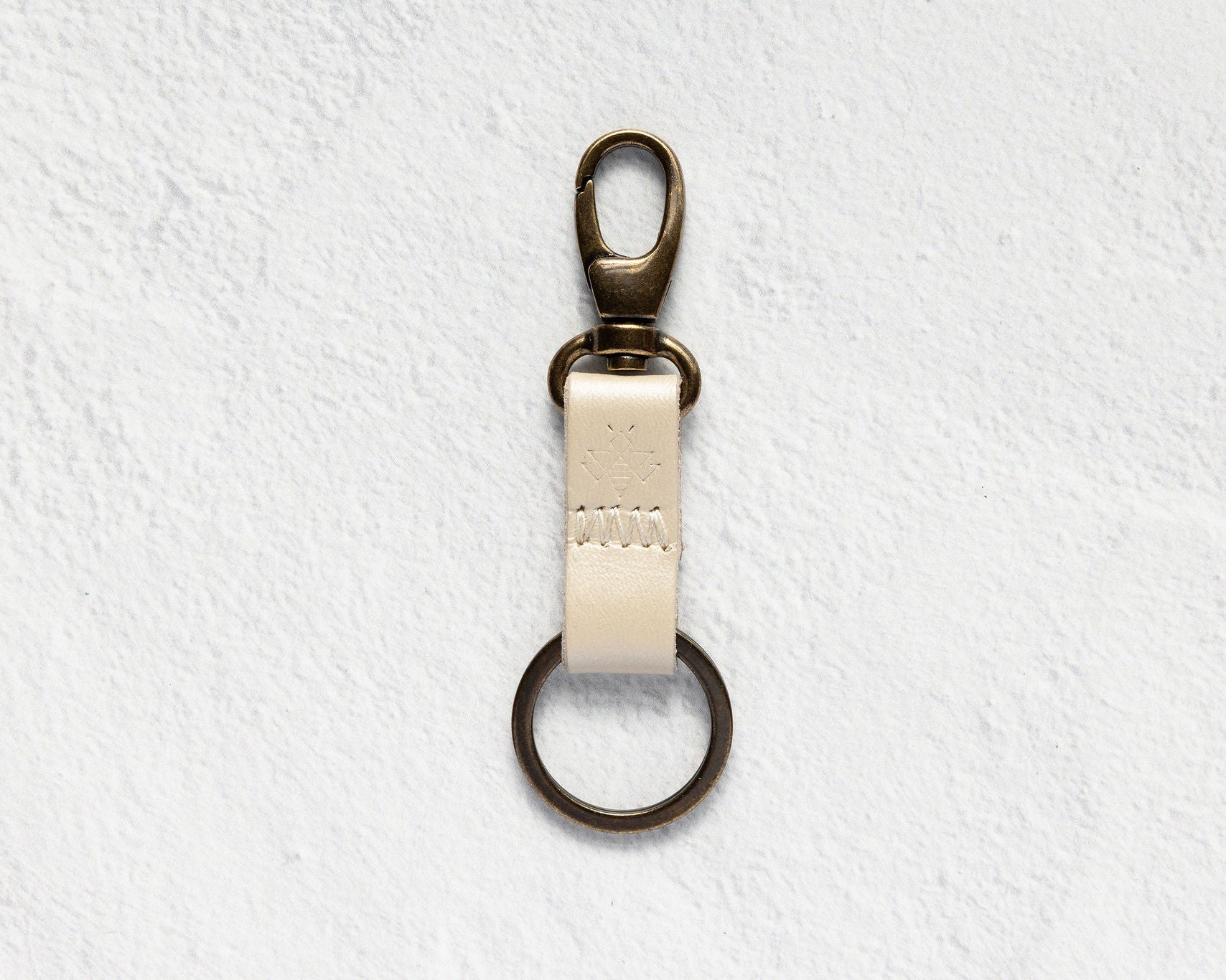 A brass key ring clasp with a cream-colored leather strap, shown against a textured white background.