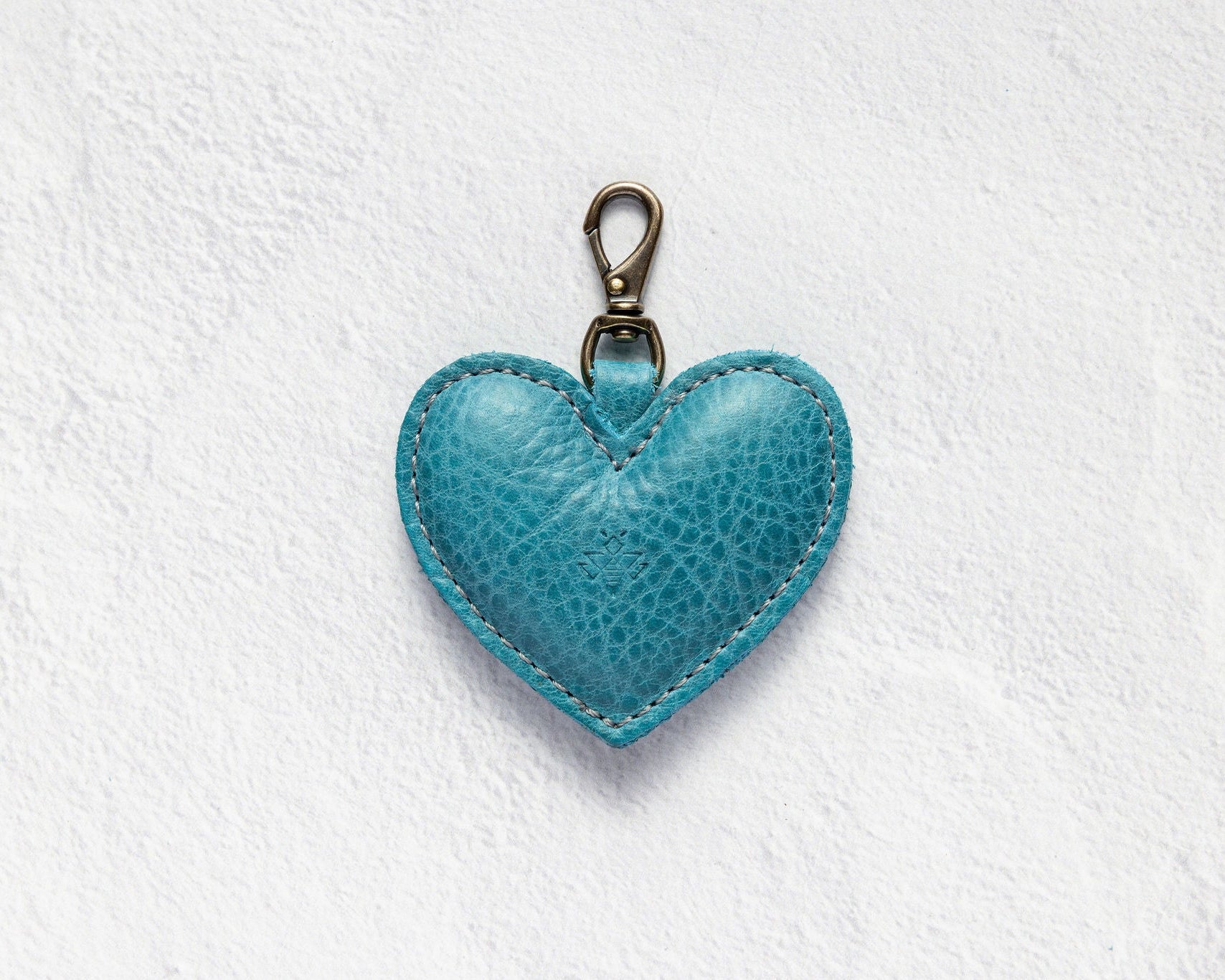 A turquoise blue textured leather heart-shaped keychain with a brass clasp, displayed on a white textured background.