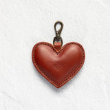 A heart-shaped brown leather keychain bag with a brass lobster clasp, centered on a textured white background.