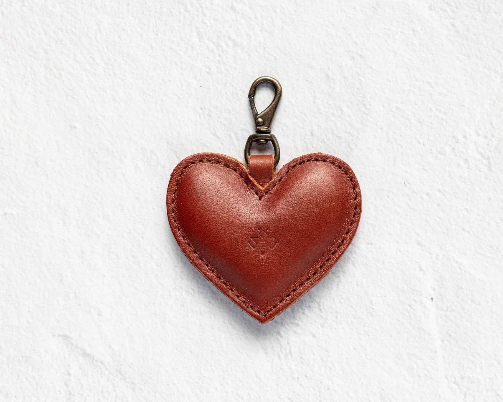 A heart-shaped brown leather keychain bag with a brass lobster clasp, centered on a textured white background.