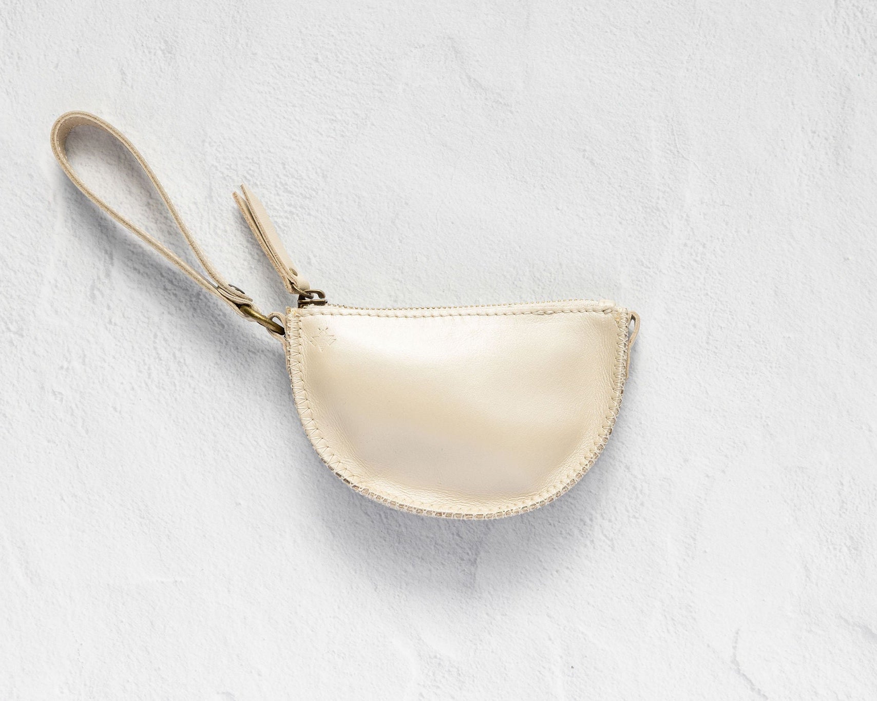 A small cream-colored leather wristlet pouch with a matching wrist strap and gold-tone zipper, displayed on a textured white background.