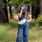 A woman stands in a wooded outdoor setting wearing a blue denim jumpsuit, a wide-brimmed dark hat, and a white crossbody bag.