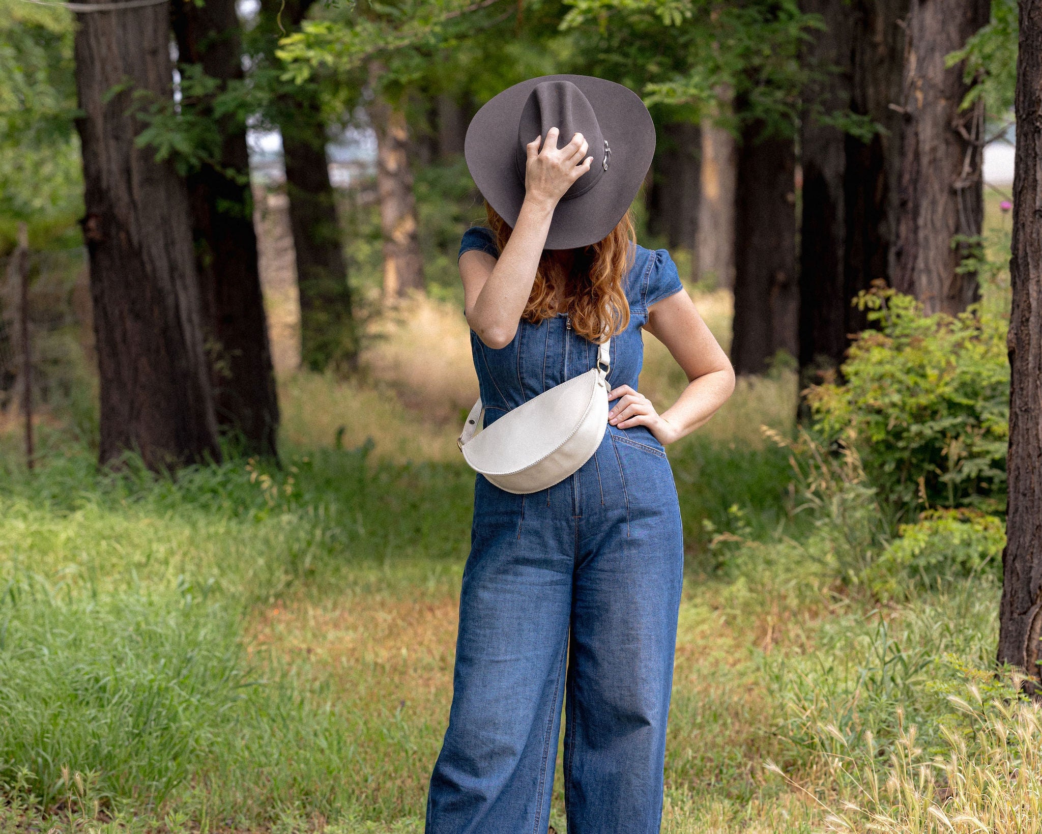 A woman stands in a wooded outdoor setting wearing a blue denim jumpsuit, a wide-brimmed dark hat, and a white crossbody bag.