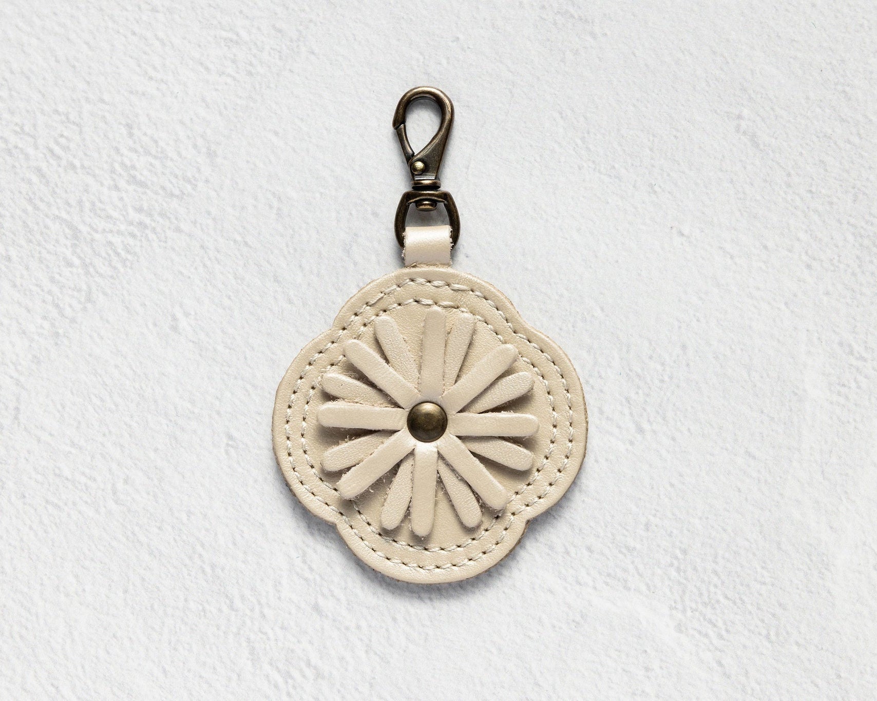 A cream-colored leather keychain or bag charm with a stitched daisy flower design and a silver-tone metal clasp, shown against a textured white background.