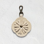 A cream-colored leather keychain or bag charm with a stitched daisy flower design and a silver-tone metal clasp, shown against a textured white background.