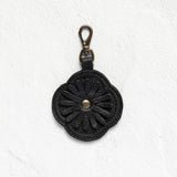 A black crocheted flower-shaped keychain or bag charm with a brass lobster clasp, displayed against a textured white background.
