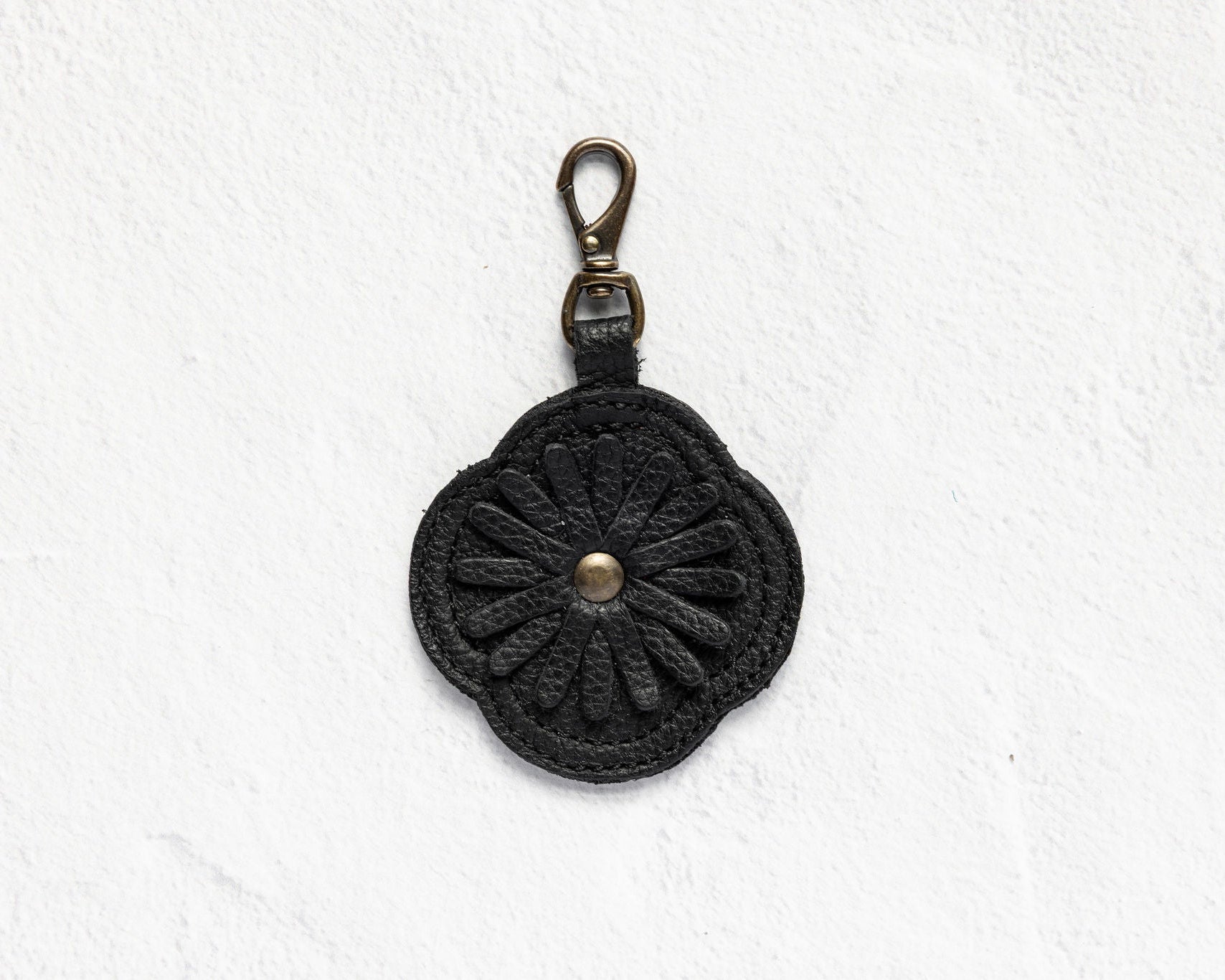 A black crocheted flower-shaped keychain or bag charm with a brass lobster clasp, displayed against a textured white background.