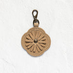 A tan leather flower-shaped keychain or bag charm with a metal clasp, displayed against a textured white wall.