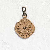 A tan leather flower-shaped keychain or bag charm with a metal clasp, displayed against a textured white wall.