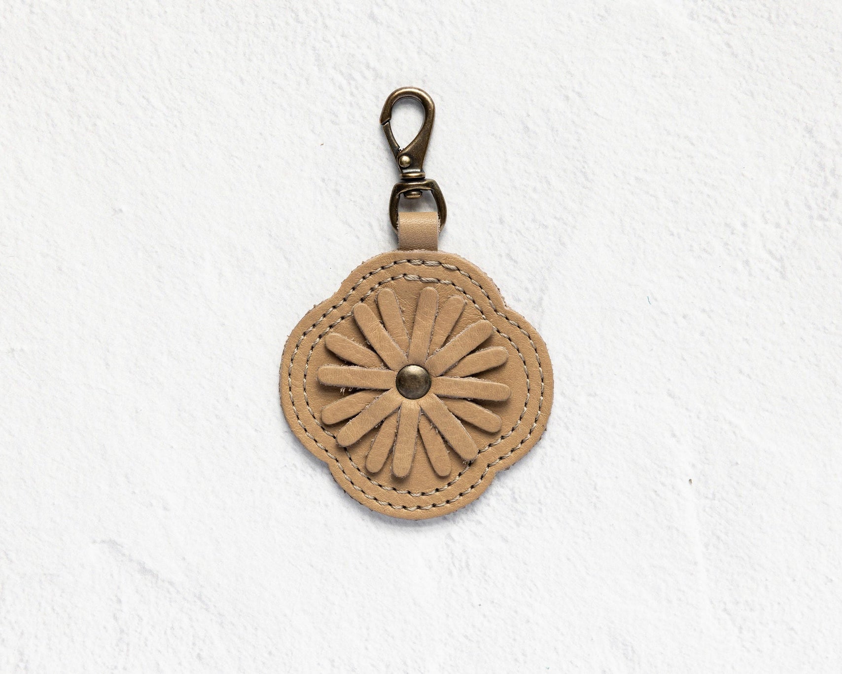 A tan leather flower-shaped keychain or bag charm with a metal clasp, displayed against a textured white wall.