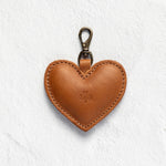 A heart-shaped brown leather keychain or bag charm with a brass-colored lobster clasp, shown against a textured white background.