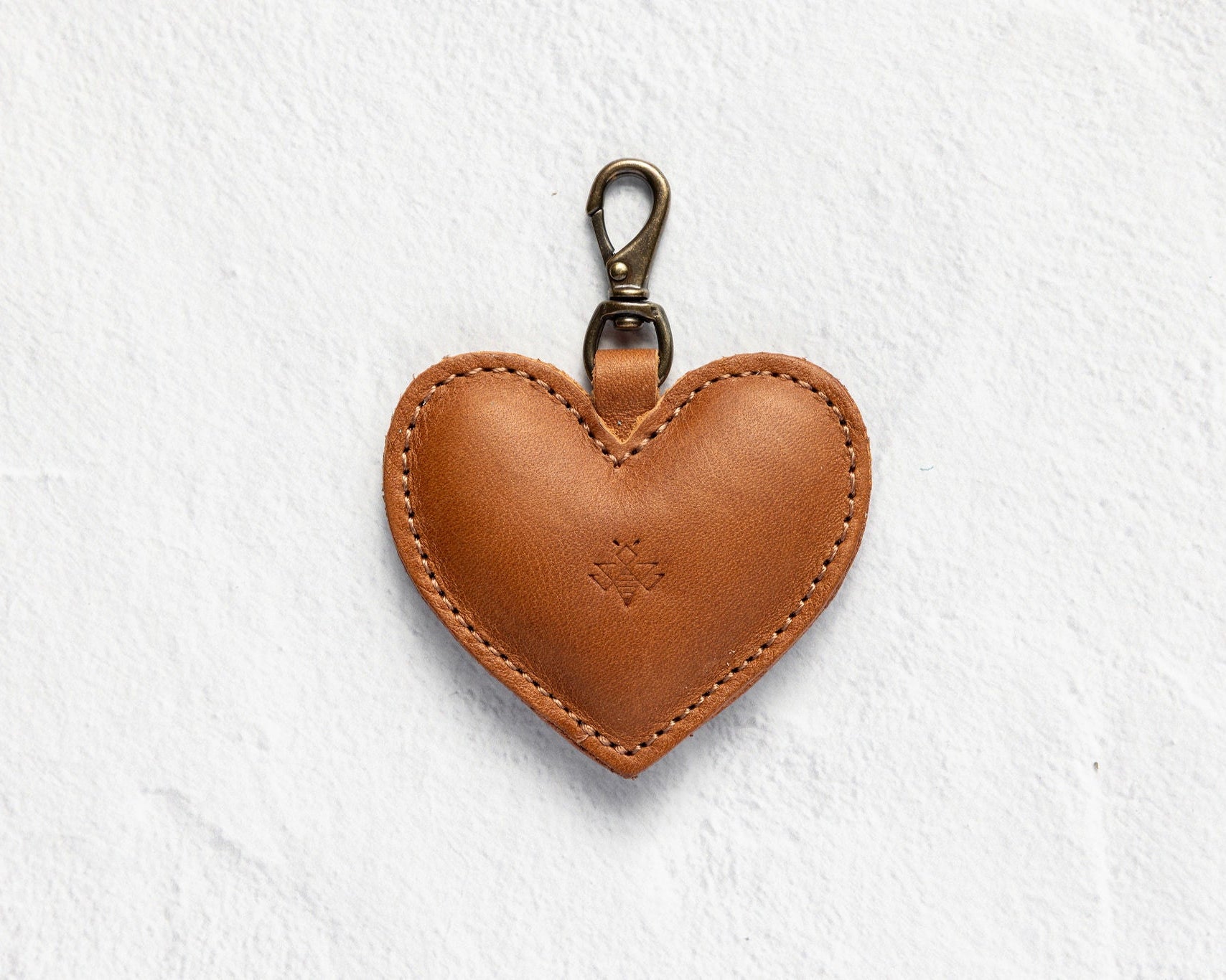 A heart-shaped brown leather keychain or bag charm with a brass-colored lobster clasp, shown against a textured white background.