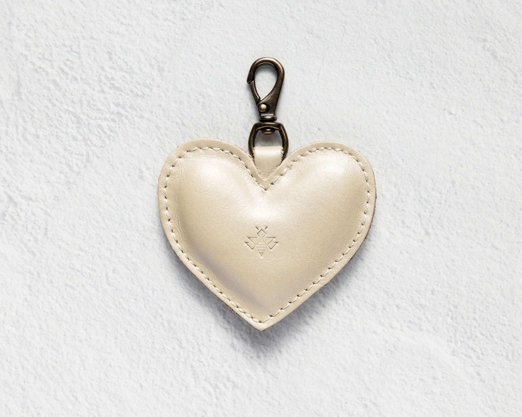 A cream-colored leather heart-shaped keychain with a silver-tone lobster clasp, displayed on a textured white background.