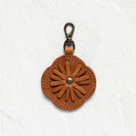 A brown leather flower-shaped keychain or bag charm with a metal lobster clasp, displayed against a textured white wall.
