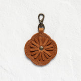 A brown leather flower-shaped keychain or bag charm with a metal lobster clasp, displayed against a textured white wall.