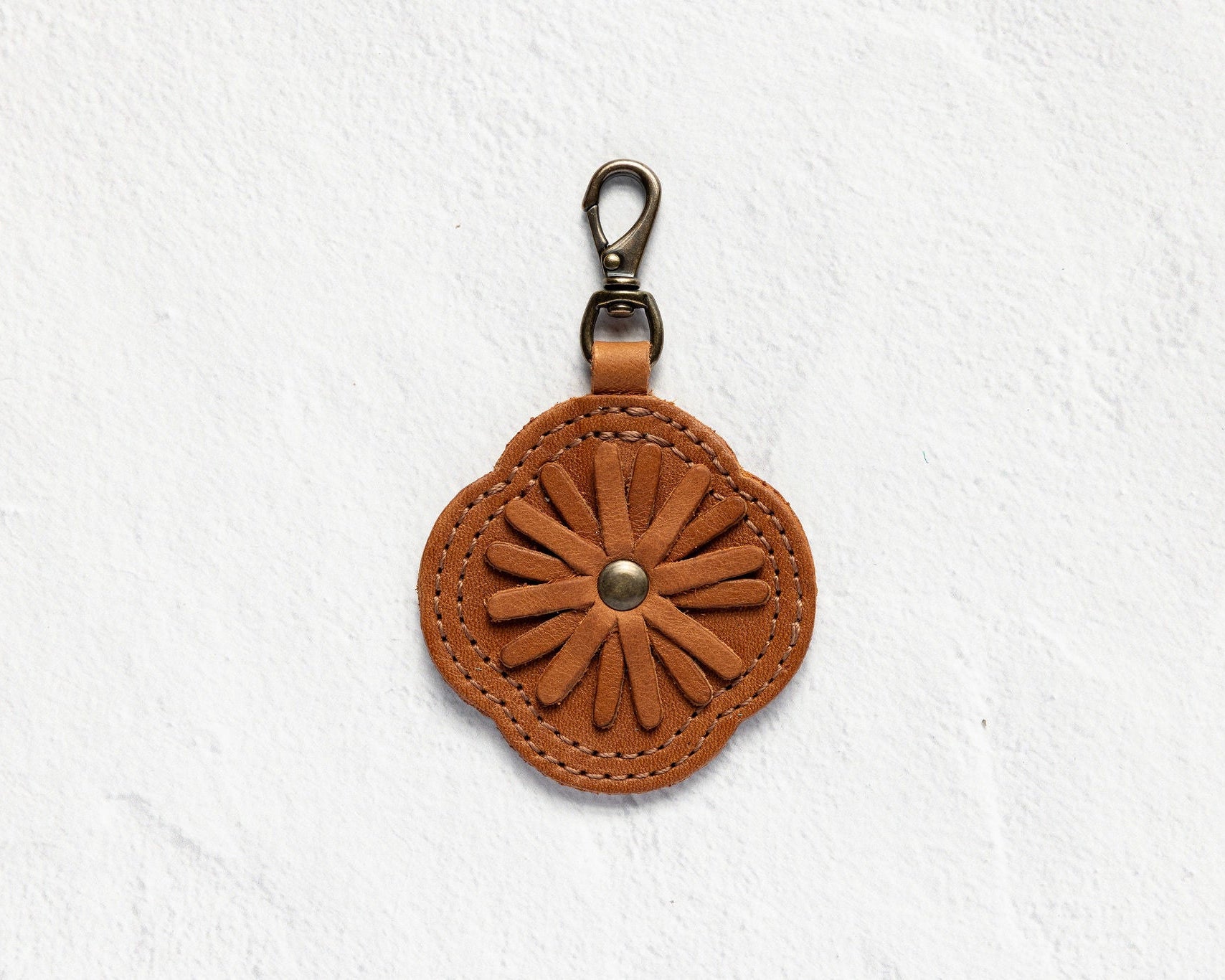 A brown leather flower-shaped keychain or bag charm with a metal lobster clasp, displayed against a textured white wall.