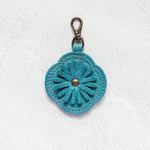 A turquoise blue flower-shaped leather keychain or bag charm with a metal clasp, displayed on a textured white background.