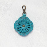 A turquoise blue flower-shaped leather keychain or bag charm with a metal clasp, displayed on a textured white background.