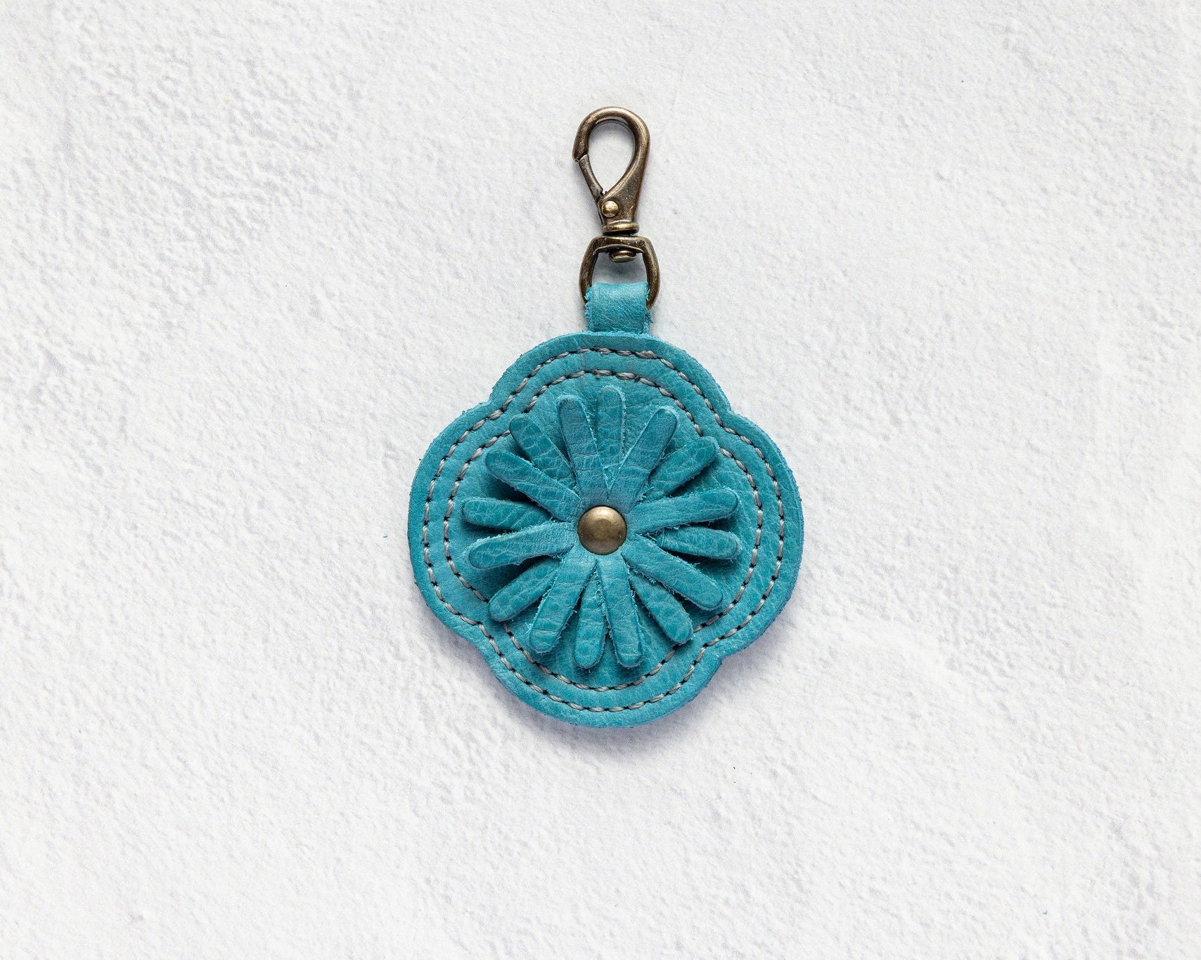 A turquoise blue flower-shaped leather keychain or bag charm with a metal clasp, displayed on a textured white background.