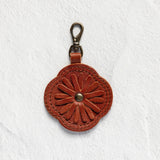 A small brown leather keychain or bag charm with an embossed floral pattern and a metal clasp, shown against a textured white background.