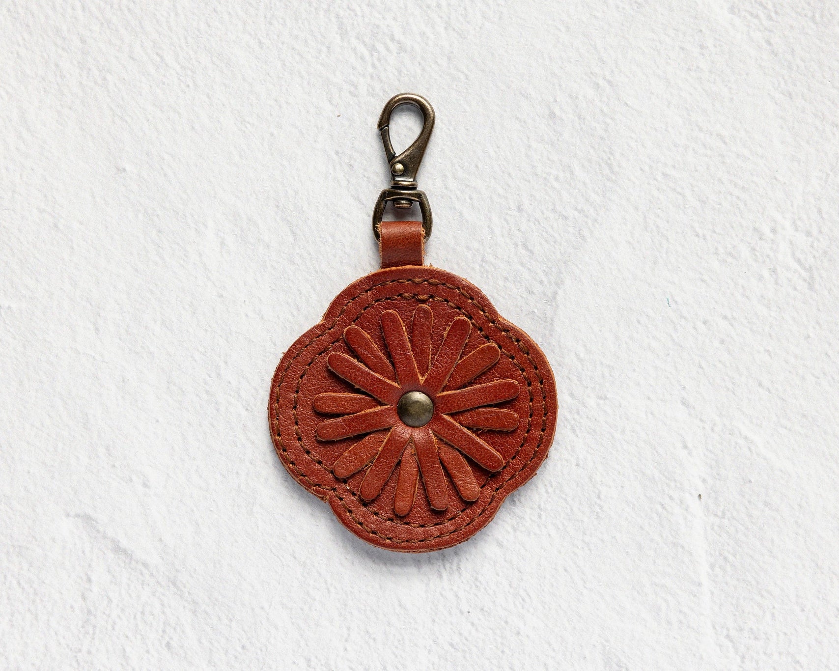 A small brown leather keychain or bag charm with an embossed floral pattern and a metal clasp, shown against a textured white background.