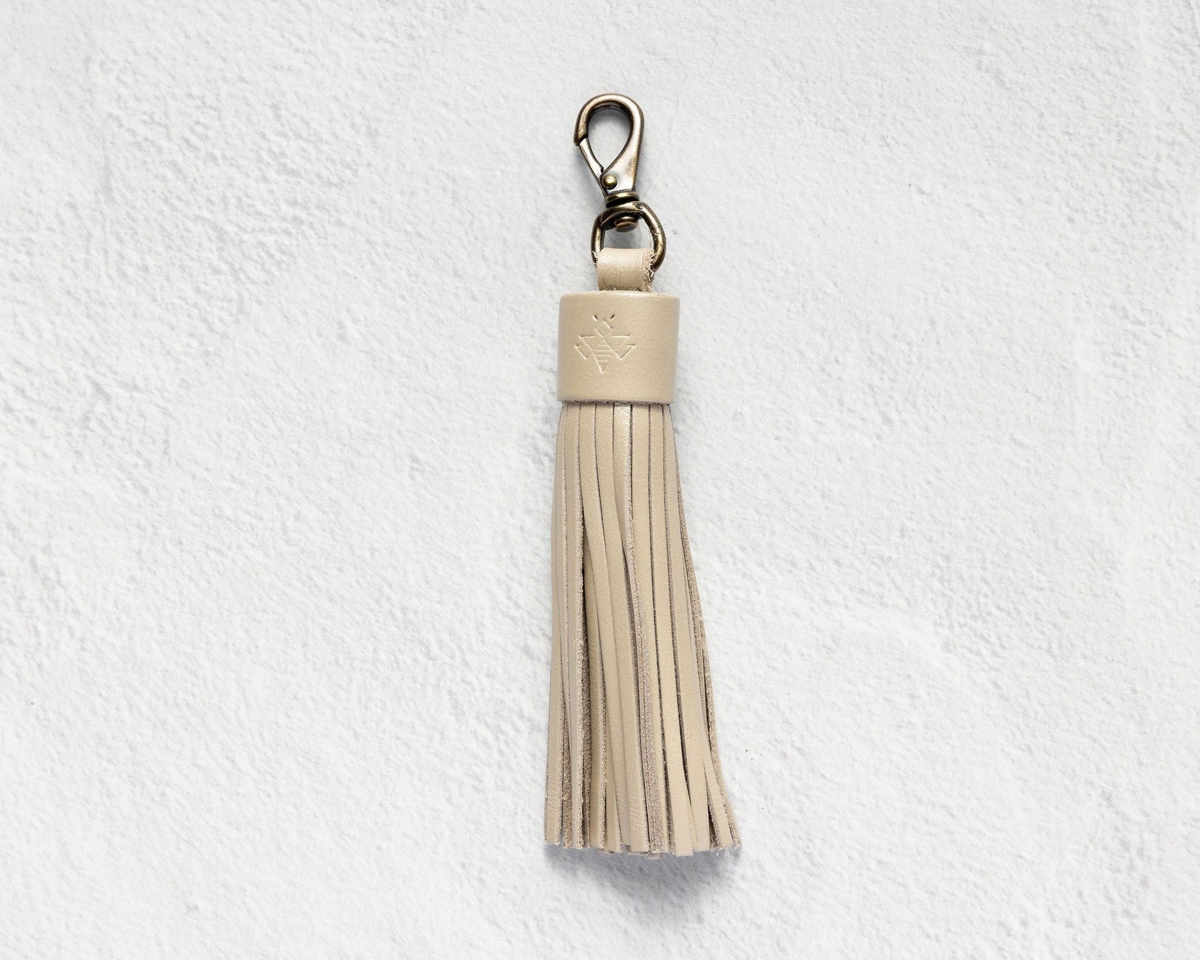 A light beige leather tassel keychain with a silver-tone lobster clasp, shown against a textured white wall.