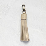 A light beige leather tassel keychain with a silver-tone lobster clasp, shown against a textured white wall.
