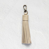 A light beige leather tassel keychain with a silver-tone lobster clasp, shown against a textured white wall.