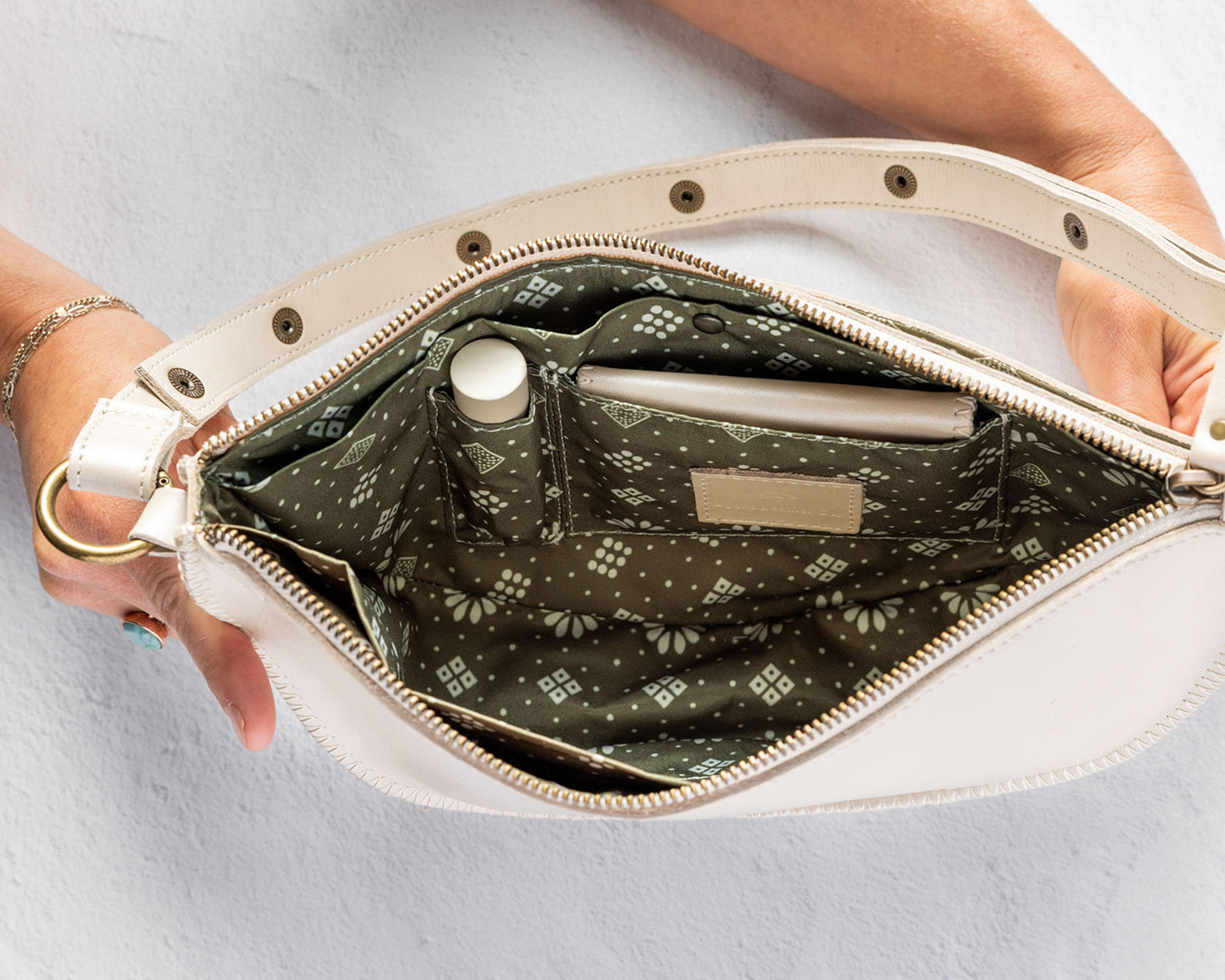 An open cream-colored shoulder bag with a patterned olive green interior, held open by two hands against a light gray background.